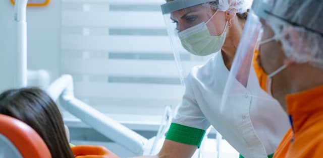 Two patients in PPE treat a patient in a dental office.