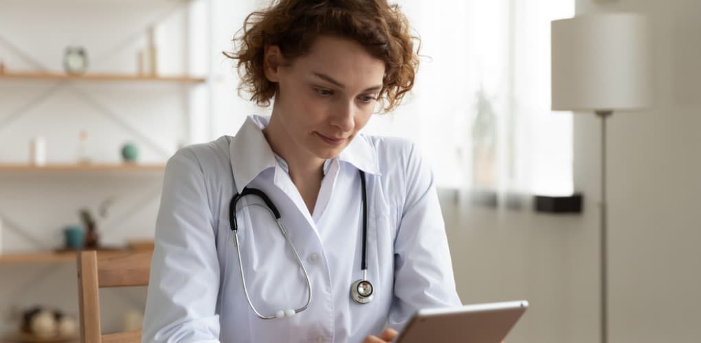Woman in a white lab coat and stethoscope around her neck looks at a tablet screen