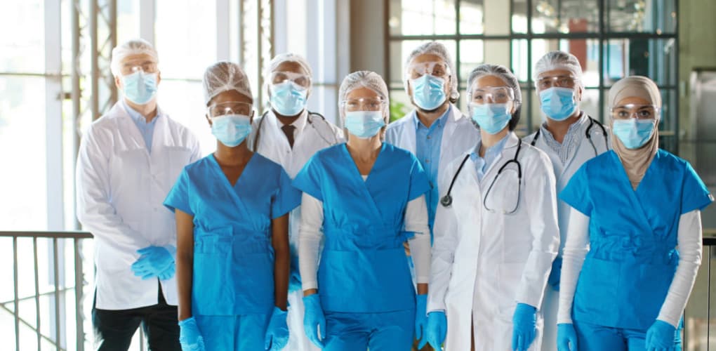 Several people in white coats and blue scrubs are wearing blue medical masks staring at the camera.