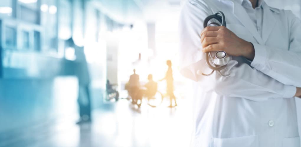 A doctor in a white lab coat is holding a stethoscope and is posing in a hospital hallway.
