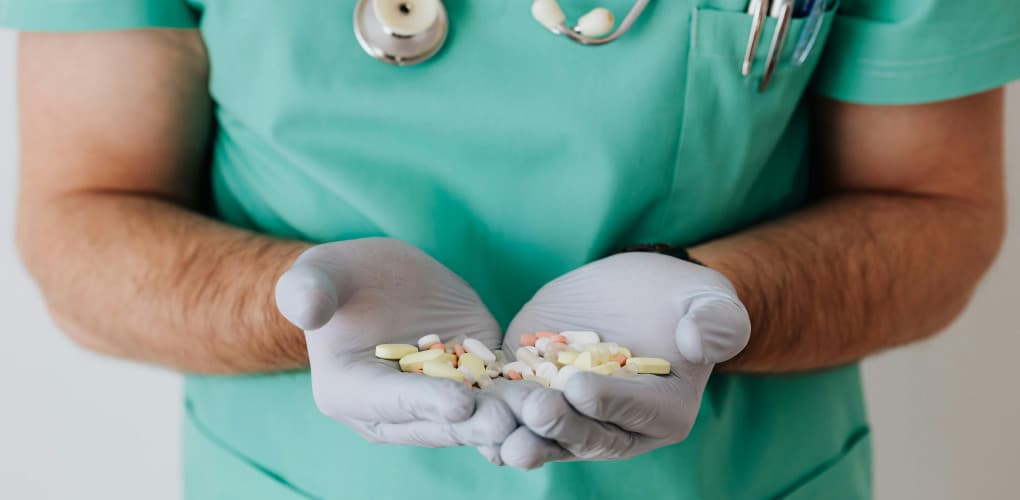 close-up of the hands of a medical professional who is wearing green scrubs and white gloves. their hands are full of pills.