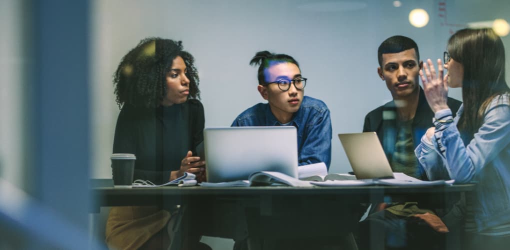 Four young adults with laptops and textbooks open talk in a conference room