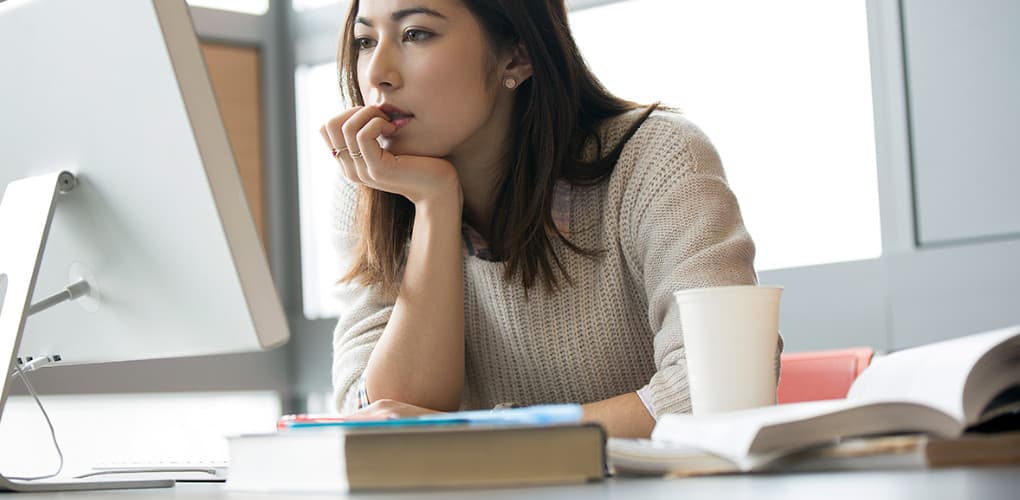 Woman in a tan sweater sits at her desk and watches a webinar on her computer monitor.