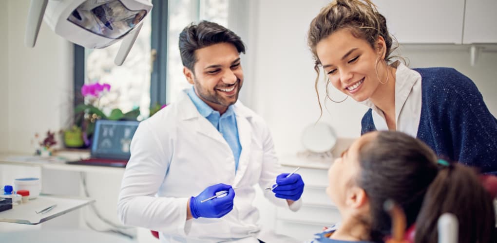 A male dentist in a white coat discusses treatment procedures with a young patient and the patient's mom.