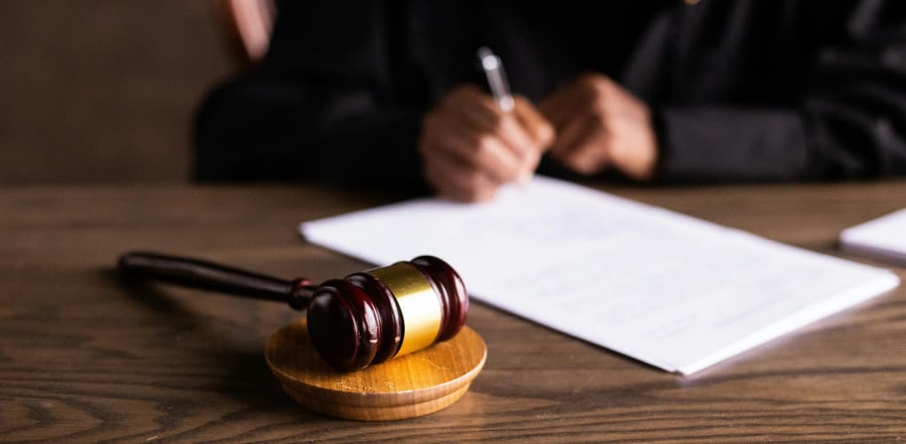 close-up of a desk with a judge's gavel in the foreground and a pair of hands in background signing a document with a pen