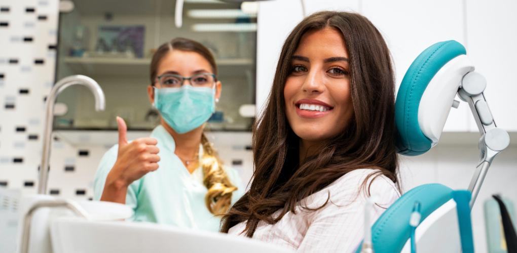 A female dentist wearing a mask gives a thumbs up behind a smiling female patient.