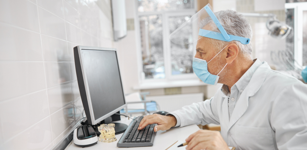 May Webinars blog — image description: A male dentist in PPE uses a computer in his office.