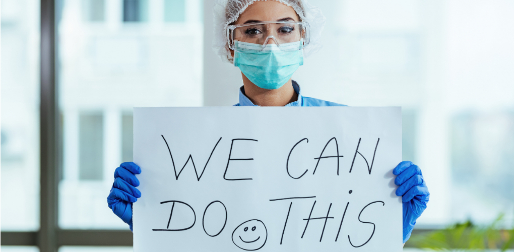 Female dental professional wearing PPE holds up a hand written sign reading 