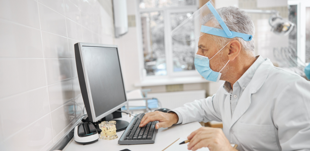 A older male dentist wearing a face mask sits at a desk and uses a computer in a dental office.