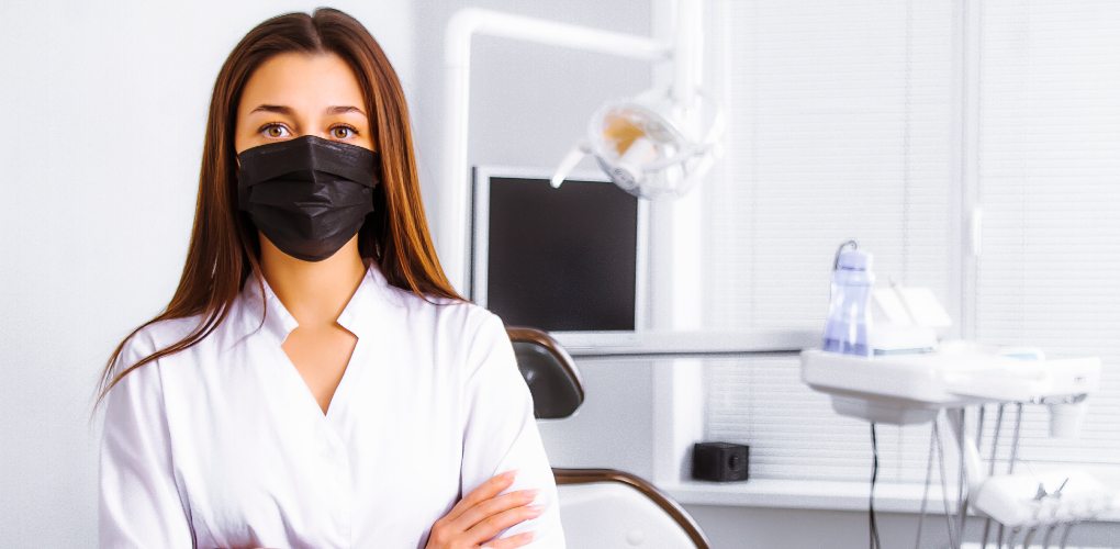 Female dentist wearing a black mask stands with her arms crossed in an empty dental exam room